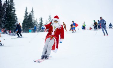 Bukovel, Ukrayna - 09 Aralık 2018: Kar maskeli kayakçı kırmızı maskeli Noel Baba kostümü giymiş beyaz sakallı kayakçı gri gökyüzünün altında karlı yamacın tepesinden diğer kayakçılarla birlikte kayak yapıyor.