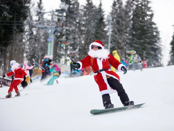Bukovel, Ukrayna - 09 Aralık 2018: Noel Baba kostümlü snowboardcu elinde selfie monopod olan beyaz sakallı, kar kayağı yarışmasında snowboard 'a biniyor.