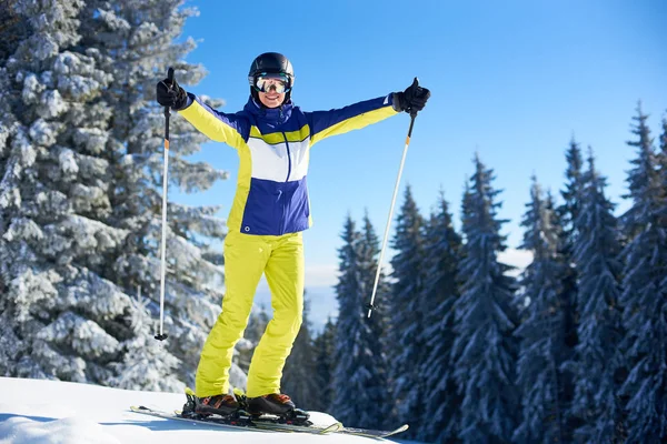 在阳光明媚的冬日 年轻的女性穿着滑雪服摆姿势 滑雪者手拿着滑雪杆 高高兴兴地竖起大拇指 寒假愉快地度过 背景上茂密的针叶林 — 图库照片