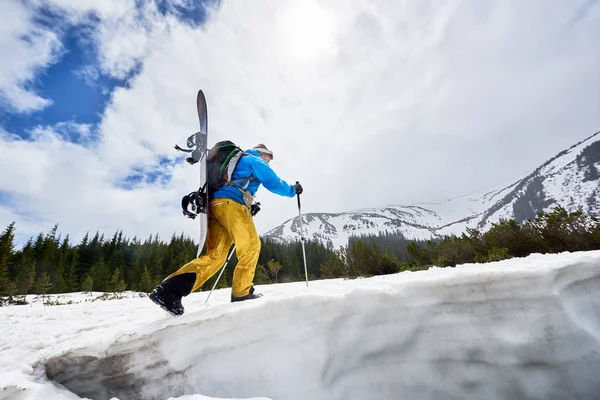 Mountaineer during backcountry skiing walking up along snowy ridge with snowboard and backpack on his back