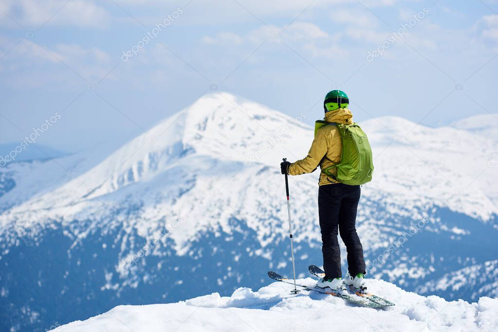 Sportsman skier in helmet and goggles with backpack riding down steep snowy slope on copy space background of blue sky and beautiful mountain landscape
