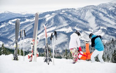Canlı kış kıyafetleri giyen bir grup arkadaş dağlarda kar altında eğleniyorlar. Kayak direkleri ve kayaklar karda sıkıştı, insanlar karda oynuyor. Kış eğlencesi