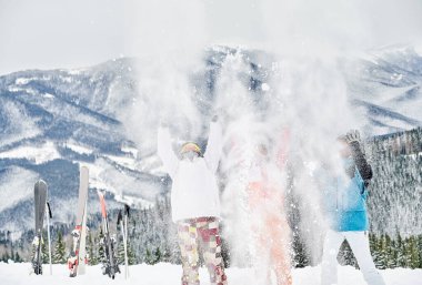 Kayak takımlı üç kayakçı havaya taze toz kar fırlatıyor, arka planda güzel dağlar olan kayak merkezinde eğleniyorlar. Kış sporları, eğlence ve ilişkiler kavramı.