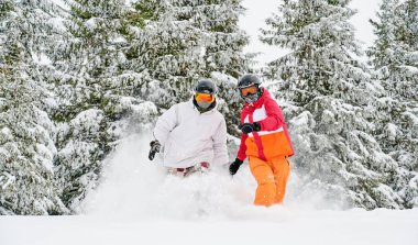Kayak takımları ve kasklı kadın ve erkek kayak merkezinde birlikte vakit geçiriyorlar, kış ormanlarında kar taneleri arasında koşuyorlar ve arka planda karlı kozalaklı ağaçlar var. Aktif boş zaman kavramı.
