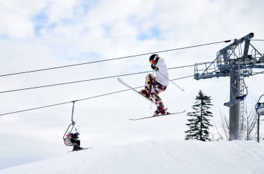 Arka planda bulutlu gökyüzü ve kayak kaldırmalarıyla havada zıplayan kayakçının yan görüntüsü. Kayak yapan bir erkek kayakçı, karla kaplı yamaçlardan kayarken numaralar yapıyor. Aşırı kış sporları kavramı