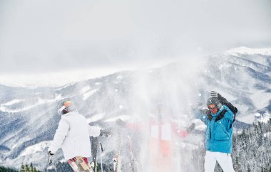 Kayak takımlarıyla kayak yapan arkadaşlar havaya taze toz kar fırlatıyorlar. İki erkek ve kadın arka planda güzel dağlarla karlı tepede eğleniyorlar. Kış eğlencesi, dostluk kavramı.