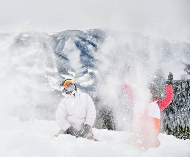 Arka planda büyüleyici manzaralara karşı karda oynayan iki genç kayakçı kar fırlatıyor. Beyaz kar üzerinde canlı renkli takım elbiseler. Kış sporları, eğlence ve ilişkiler kavramı.
