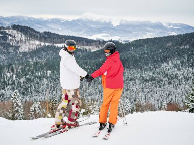 Erkek ve kadın kayakçılar el ele tutuşup kar kaplı tepede dikilirken kameraya bakıyorlar. Genç çift kış günü dağlarda birlikte kayak yapıyor. Kış sporları ve ilişkiler kavramı
