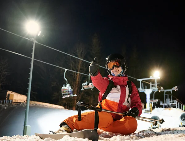 夜にはライトアップされたリフトの下で雪に覆われたスキー場に座って鮮やかなスーツを着ている女の子のスキーヤーは 親指を表示幸せとスキーリゾートでのフリーライド後に満足しています 極端なスポーツの概念 — ストック写真