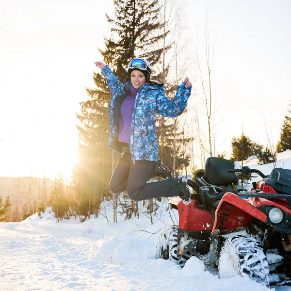 美しいブロンドの女の子の肖像画 青い冬のジャケットを着て雪の山の中で冬には赤の4輪Atvの横にジャンプ 背景に明るい夕日 アクティブライフスタイルコンセプト — ストック写真