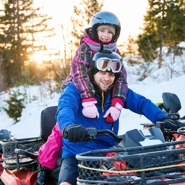 Portrait of smiling family, father and daughter, wearing vivid ski costumes, helmets and goggles, sitting on squad-bike, smiling to camera in snowy mountains on sunset. Concept of active leisure.