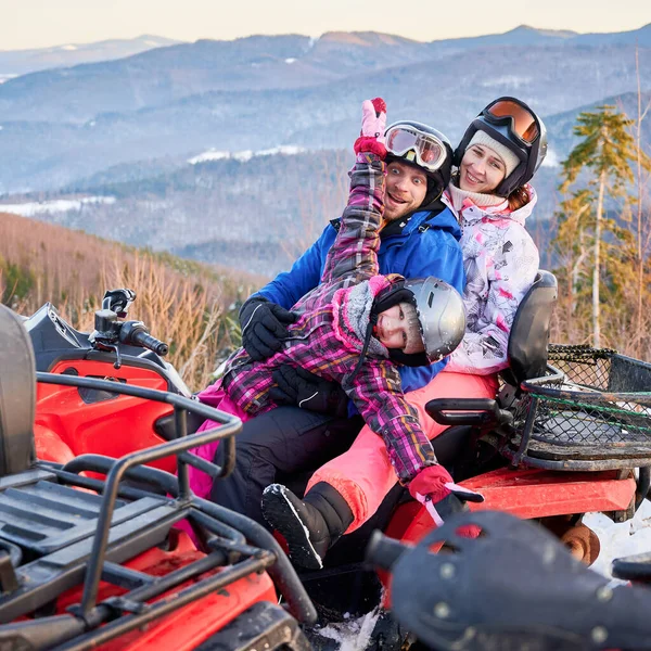 3つの幸せな家族は カラフルなスキースーツを着て 素晴らしい冬の休日を過ごし 赤いクワッドバイク サイドビュー 背景に山の尾根に雪の山の中で一緒に抱き合って — ストック写真