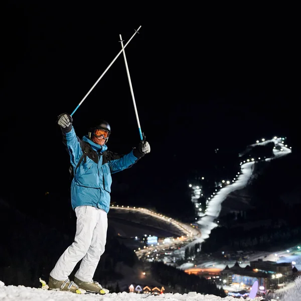 Male skier in winter ski jacket and helmet raising ski poles and looking at camera. Young man in ski goggles standing on snow-covered slope with night ski resort on background. Concept of night skiing