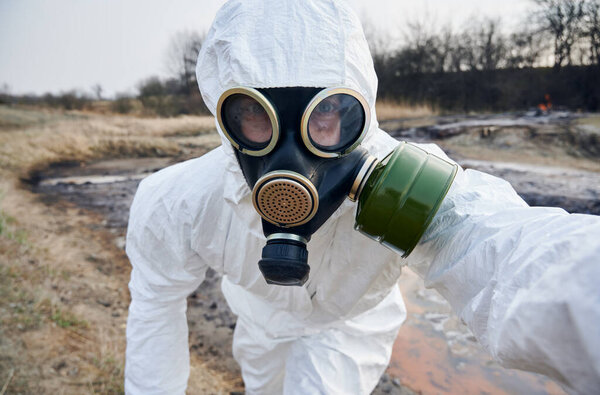 Close-up snapshot of scientist in gas mask and coverall looking right at camera, working on contaminated territory , rusted water on background. Concept of polluted nature