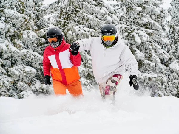 若いカップルの観光客は 背景に愛らしいスプルースの森に対して雪の中で楽しく落下します 白い雪の上に鮮やかなカラフルなスーツ 冬のヨーロッパスキーリゾート — ストック写真