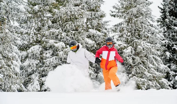 背景に雪深い針葉樹の冬の森の中でパウダースノーを駆け抜けるスキースーツやヘルメットの女性と男性 ゴーグルの若いスキーヤーはスキー場で一緒に時間を過ごす — ストック写真