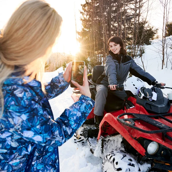 友人がスマートフォンで写真を撮っている間 美しいブルネットの女性はすべての地形の車に座って笑顔 冬の山の中で晴れた日にクワッドバイクで若い女性 クワッドバイクの概念 — ストック写真