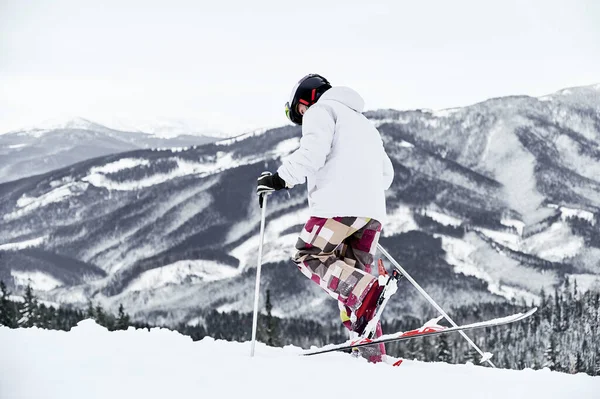 Back view of alpine skier in jacket and helmet skiing in mountains. Man freerider with ski sticks walking through the snow with beautiful snowy hills on background. Concept of winter sport activities.