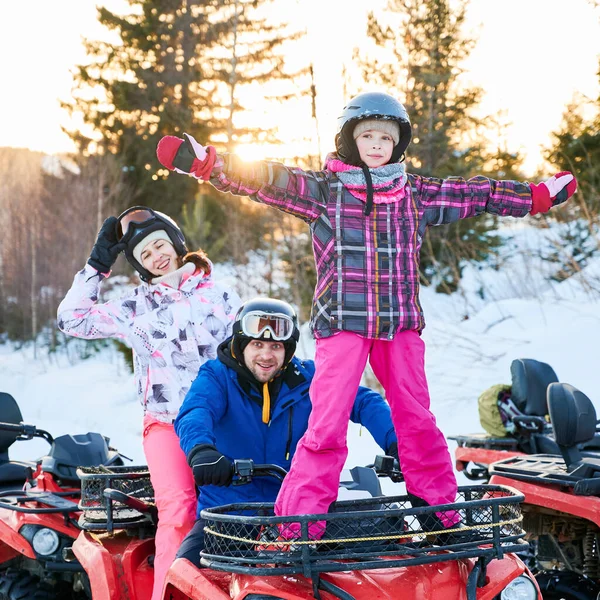 Young family in helmets smiling to camera while sitting on all-terrain vehicle with cute daughter. Adorable little girl in winter jacket enjoying quad biking with parents. Concept active leisure.