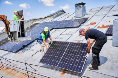 İşçiler vinç kaldırma yardımıyla evin metal çatısına güneş paneli inşa ediyorlar. Dışarıda fotovoltaik güneş modülü taşıyan iki adam. Yenilenebilir enerji üretimi kavramı.