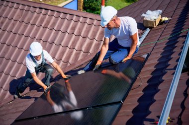 İşçiler evin çatısına güneş paneli inşa ediyorlar. Dışarıda fotovoltaik güneş modülü kuran kask takan iki adam. Alternatif, yeşil ve yenilenebilir enerji üretimi kavramı.