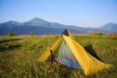 Yellow tourist tent on grassy meadow, surrounded by wildflowers, with majestic mountains in background. Clear blue sky and sunlight create vibrant and peaceful camping scene in nature.