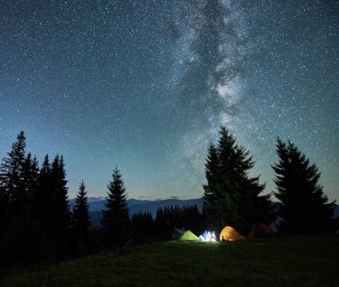 Night starry sky over grassy hill with illuminated tourist tents and coniferous trees. Mountain valley with camp tents under sky with stars and Milky Way. Hiking, night camping and astronomy concept.