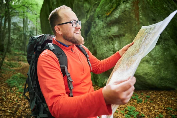 Sakallı turist yakın plan haritası incelerken sık ormandaki büyük kayaların önünde duruyor. Gri sırt çantası ve gözlüklü gezgin, açık hava macerası, yürüyüş ya da tırmanma rotası planlıyor..