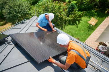 İşçiler evin çatısına güneş paneli inşa ediyorlar. Dışarıda fotovoltaik güneş modülü kuran kask takan iki adam. Alternatif, yeşil ve yenilenebilir enerji üretimi kavramı.