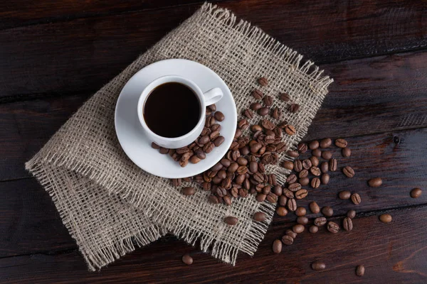 Ceașcă albă de cafea cu boabe de cafea lângă țesătura de in pe fundalul întunecat de epocă. Privire de sus . — Fotografie, imagine de stoc