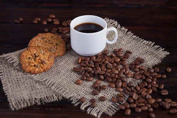 Weiße Tasse Kaffee mit Kaffeekörnern in der Nähe und Keksen auf Leinenstoff auf dunklem Vintage-Hintergrund — Stockfoto