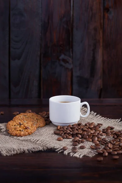 Xícara branca de café com grãos de café perto e biscoitos em tecido de linho no fundo escuro do vintage — Fotografia de Stock