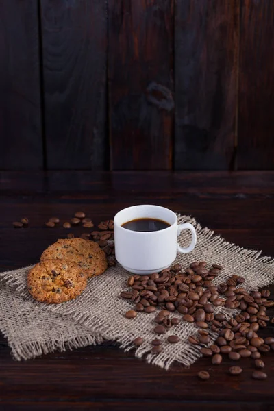 Weiße Tasse Kaffee mit Kaffeekörnern in der Nähe und Keksen auf Leinenstoff auf dunklem Vintage-Hintergrund — Stockfoto