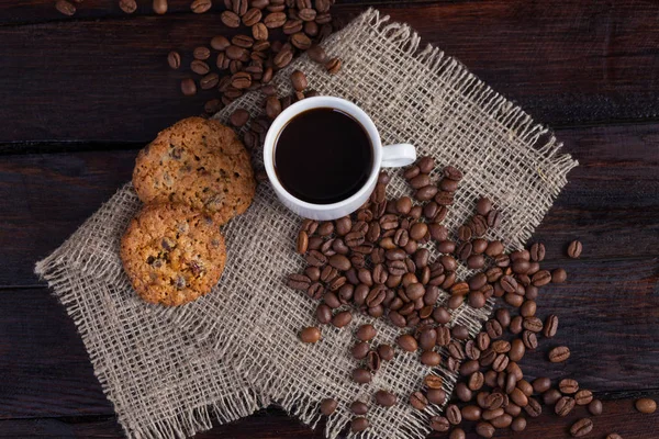 Xícara branca de café com grãos de café perto e biscoitos em tecido de linho no fundo escuro do vintage — Fotografia de Stock