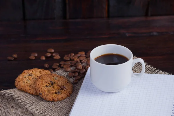 Белая чашка кофе с зерном кофе рядом и печенье и блокнот в клетке для записей на льняной ткани на темном винтажном фоне — стоковое фото