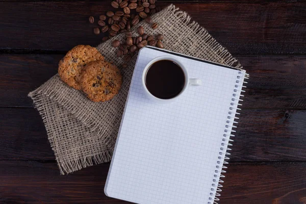 Xícara branca de café com grãos de café perto e biscoitos e bloco de notas em uma gaiola para registros em tecido de linho no fundo escuro do vintage. Vista superior — Fotografia de Stock