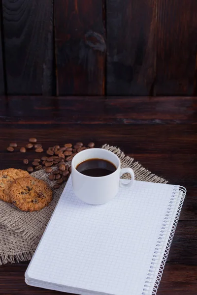 Taza blanca de café con granos de café cerca y galletas y bloc de notas en una jaula para registros sobre tela de lino en el fondo oscuro vintage. Foto vertical —  Fotos de Stock