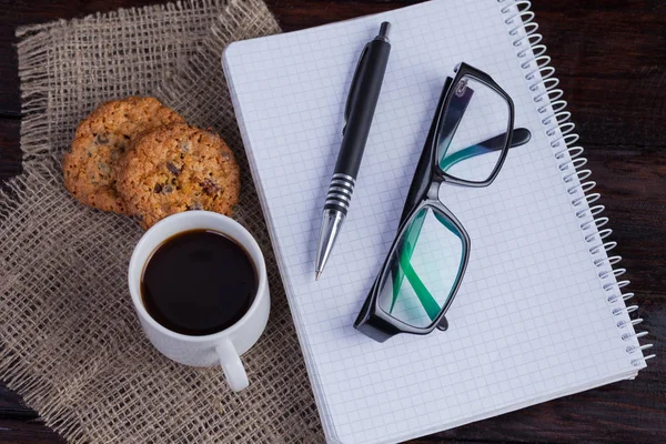 Белая чашка кофе с печеньем и блокнотом, ручка и очки на темном деревянном фоне — стоковое фото