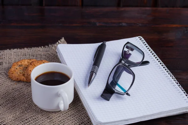 Белая чашка кофе с печеньем и блокнотом, ручка и очки на темном деревянном фоне — стоковое фото