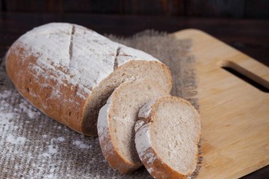 Keten kumaş parçası ile ahşap arka plan üzerinde dağınık un ile bir meşe panosuna dilimlenmiş adet taze kahverengi ekmek