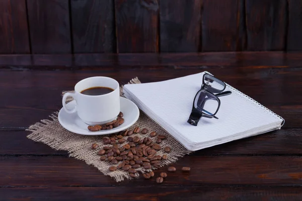 Weiße Kaffeetasse mit Notizblock und Gläsern auf dunklem Holzhintergrund und Leinenstoff — Stockfoto