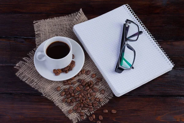 Белая чашка кофе с блокнотом и стаканами на темном деревянном фоне и льняной ткани — стоковое фото
