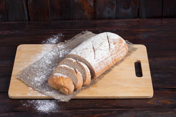 Roti coklat segar dengan potongan-potongan yang diiris di papan ek dengan tepung yang tersebar di latar belakang kayu dengan sepotong kain linen — Stok Foto