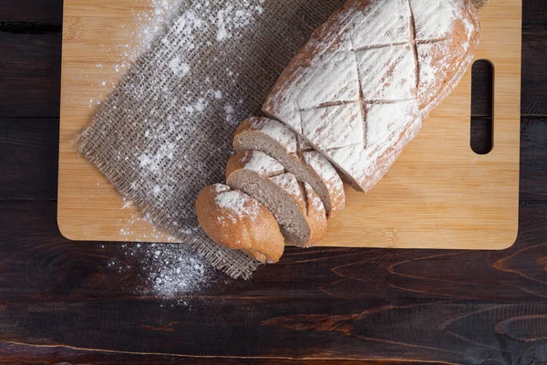 Roti coklat segar dengan potongan-potongan yang diiris di papan ek dengan tepung yang tersebar di latar belakang kayu dengan sepotong kain linen. Tampilan puncak foto — Stok Foto