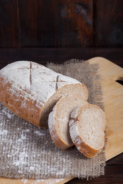 Fresh brown bread with sliced pieces on an oak board with scattered flour on a wooden background with a piece of linen fabric. Vertical photo — Stock Photo, Image