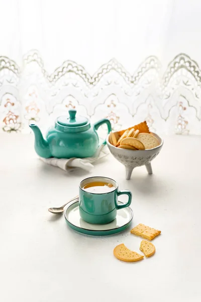 Panci teh berwarna, cangkir teh dan piring, kue di atas meja dengan jendela — Stok Foto