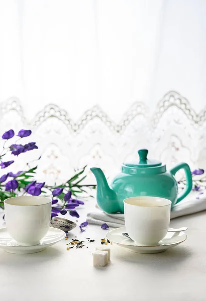 Two white tea cups with green tea, teal colored tea pot, purple flowers on white background with copy space