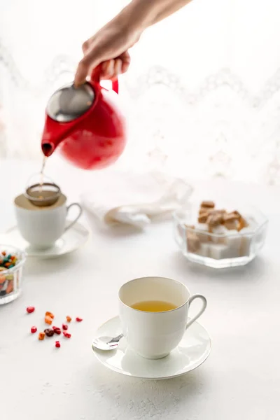 Womans Hande gieten thee uit rode theepot, witte thee bekers, Suikerpot staande op witte tafel — Stockfoto