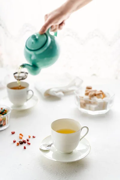 Womans hande menuangkan teh dari teko teh berwarna, cangkir teh putih, mangkuk gula berdiri di atas meja putih — Stok Foto