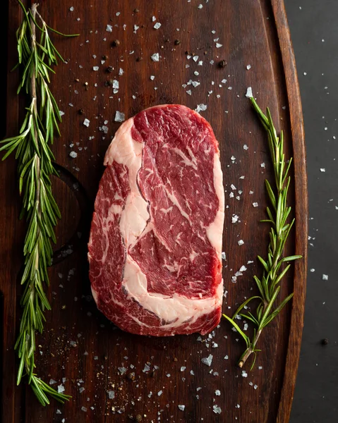 Bife de ribeye cru com alecrim fresco na placa de corte de madeira — Fotografia de Stock